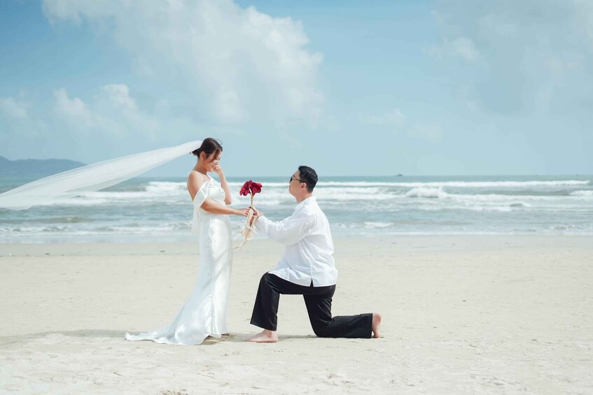 A beach setting with a person kneeling and proposing to another person holding a rose. 1