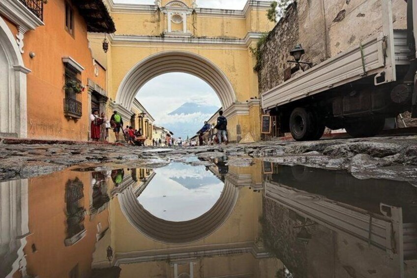 A street with a large archway, a truck parked on the right, and a puddle reflecting the surroundings. 4