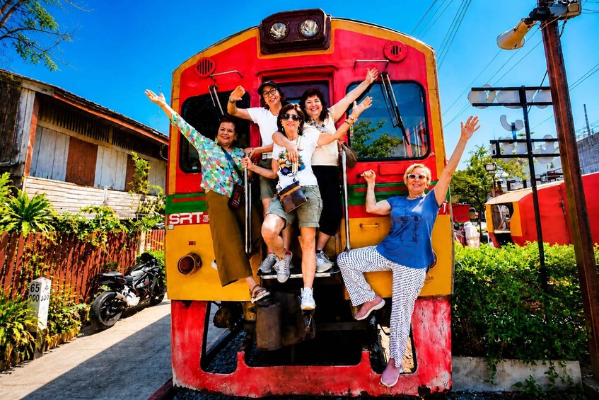 A group of people on a colorful train with a red and yellow exterior, parked on tracks with a motorcycle and a building in the background. 1