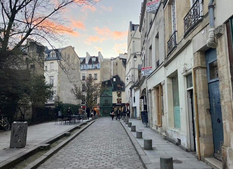 A cobblestone street in a European city with buildings on either side, some with signs indicating real estate for sale. 2