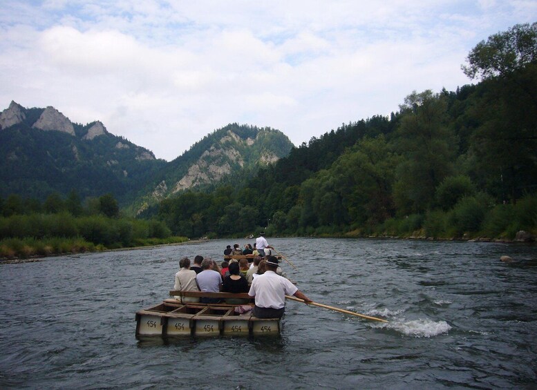 A group of people rowing a wooden boat on a river, surrounded by forested hills. 1