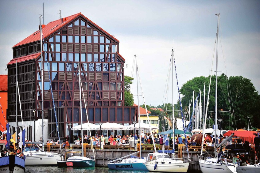 A marina with boats docked, a large building with a red roof, and people gathered near the waterfront. 1