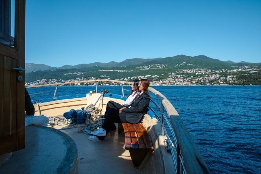 A boat with a person sitting on the deck, a clear blue sea, and a distant shoreline with buildings and hills. 1