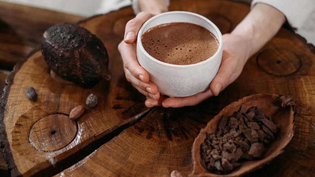 A person holding a cup of coffee with a wooden spoon and a cocoa pod on a wooden table. 1