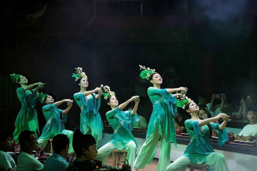 A group of dancers in teal costumes performing on stage with an audience seated in front. 5