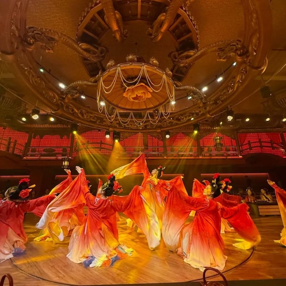 A group of dancers performing in a grand hall with ornate ceiling and red-lit backdrop. 1