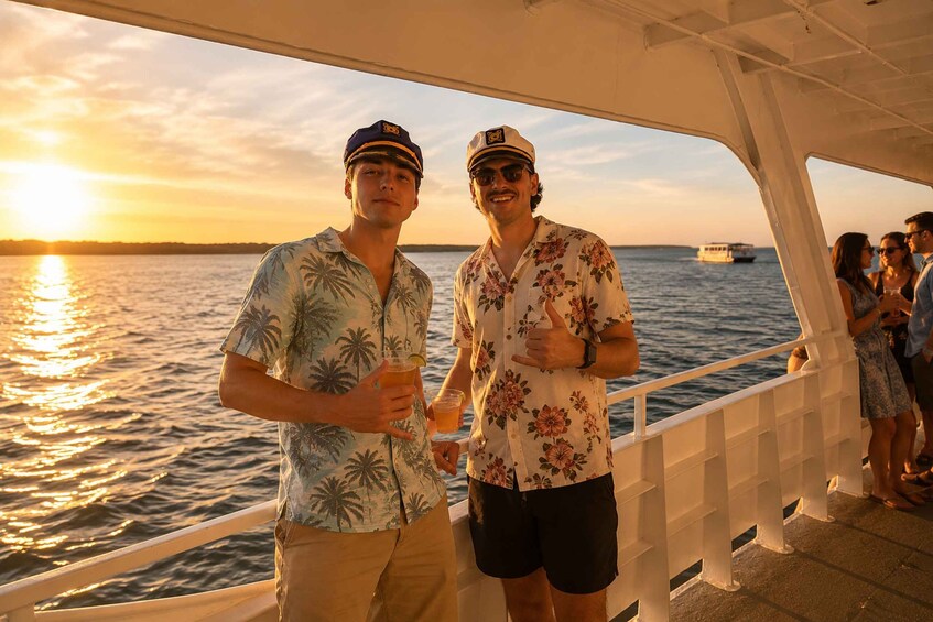Two people on a boat at sunset with drinks, a railing, and a distant boat on the water. 3