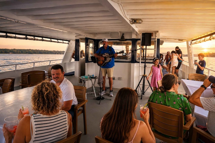 A boat with people seated at tables, a man playing guitar, and a sunset in the background. 1