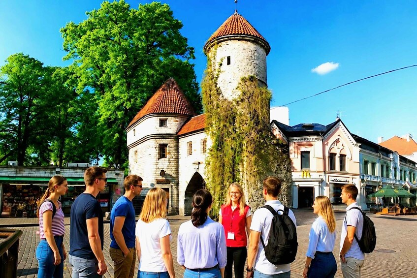 A group of people standing in front of a historic tower with a red roof, surrounded by trees and buildings with shops. 1