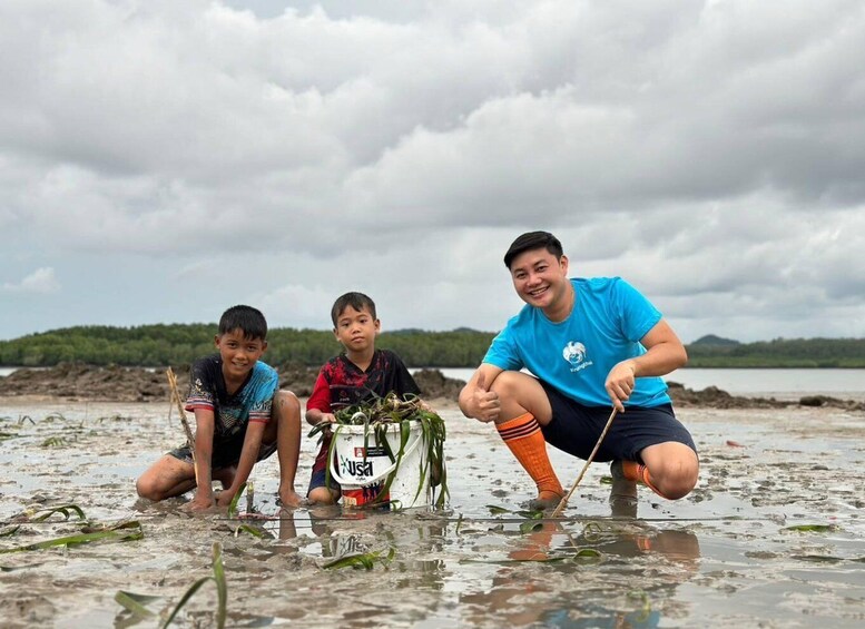 Three people collecting aquatic plants on a muddy shore with a lake and cloudy sky in the background. 1