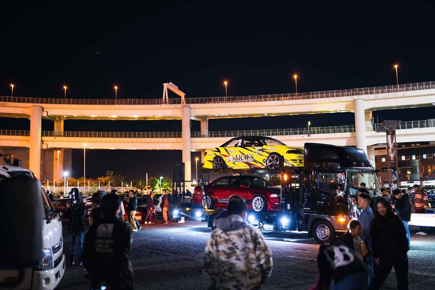 A nighttime street scene with a red car on a truck, a white van, and a crowd of people. 5