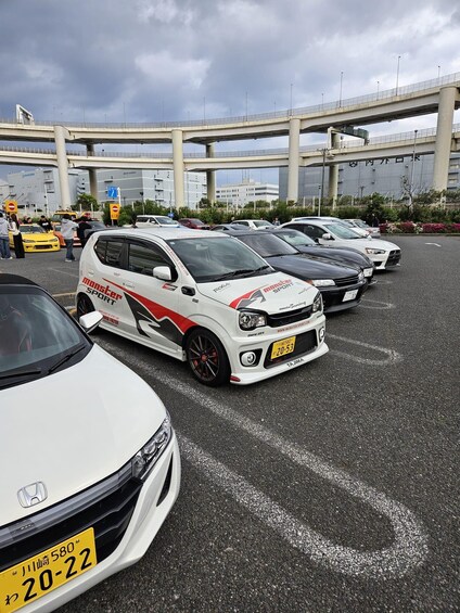 A parking lot with several modified Honda Civic cars, a bridge, and a building with signage in a foreign language. 2