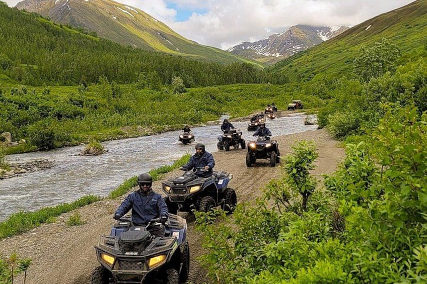 A group of off-road vehicles on a dirt path beside a river in a mountainous area. 3
