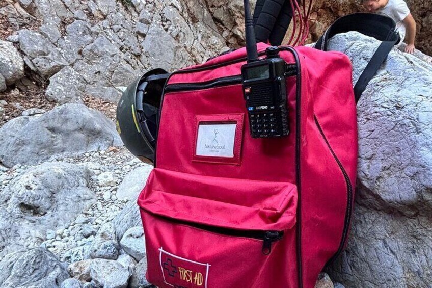 A red backpack with a first aid kit, a helmet, and a walkie-talkie on a rocky surface. 4