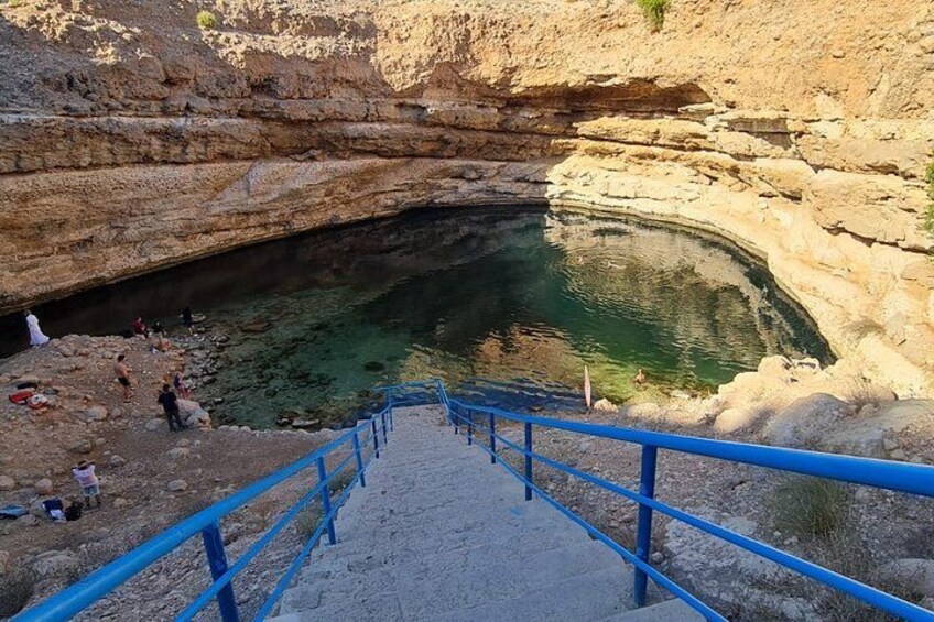 A rocky landscape with a water-filled basin, a blue staircase leading down, and people at the bottom. 2