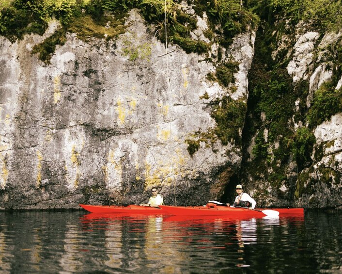 Two people kayaking in a red kayak on calm water with a rocky cliff in the background. 2