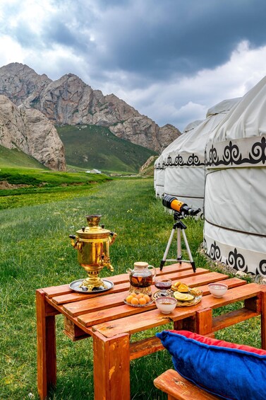 A wooden picnic table with a traditional brass samovar, bowls, and a telescope, set against a backdrop of a grassy field and mountains. 2