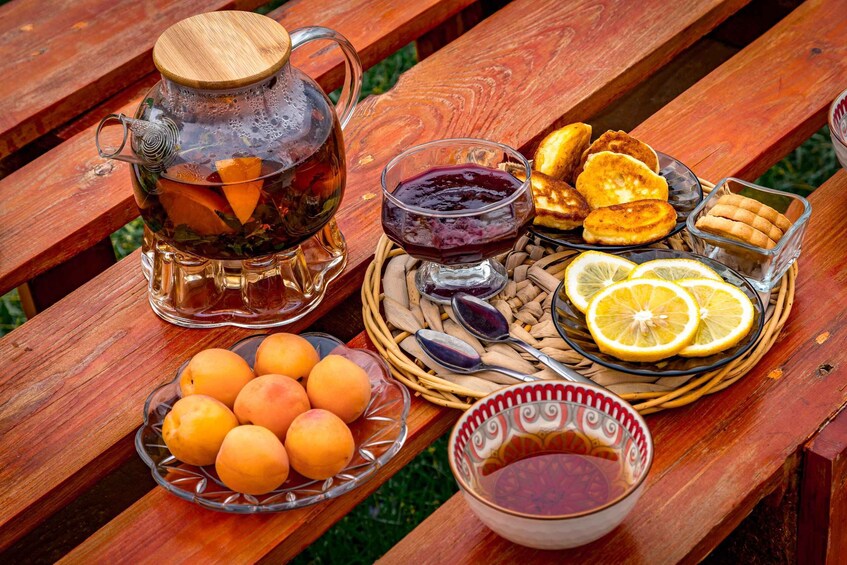 A picnic setup with a glass teapot, a bowl of apricots, a plate of bread, a glass of jam, and a bowl of honey. 5