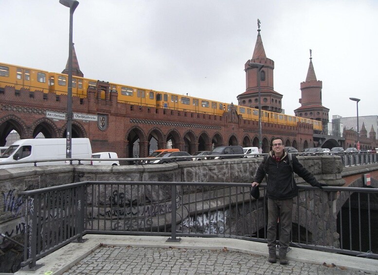 A man standing on a bridge with graffiti, a yellow tram, and a historic building with towers in the background. 4