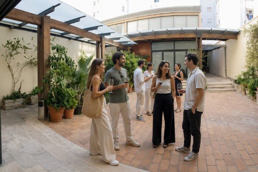 A group of people standing and conversing in a covered outdoor area with potted plants and a building in the background. 3