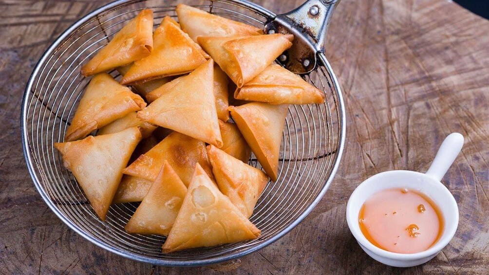 A basket of golden-brown samosas on a wire rack, accompanied by a small bowl of dipping sauce. 4