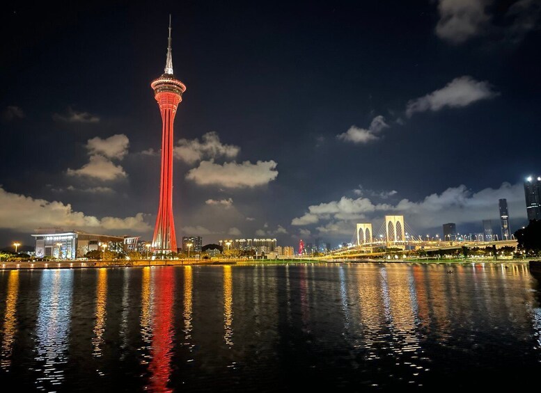 A city skyline at night with a prominent red-lit tower, a bridge, and a body of water reflecting the lights. 5