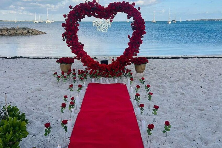 A heart-shaped floral arrangement on a beach with a red carpet leading to it. 2