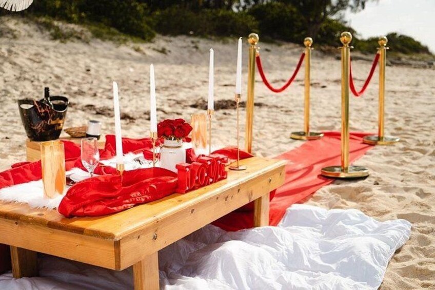 A beach picnic setup with a wooden table, red cloth, white candles, and a red rope barrier. 1