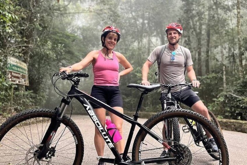 Two people on bicycles in a forested area with a paved path. 1