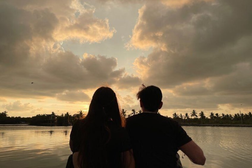 Two people sitting on a boat, facing a sunset over a body of water with palm trees in the background. 4