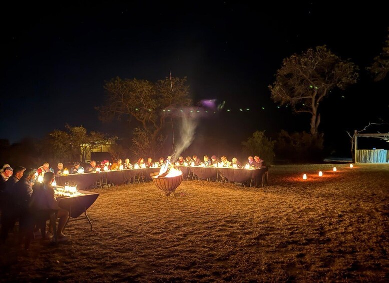 A group of people seated at a long table outdoors at night, with a fire in the center. 4