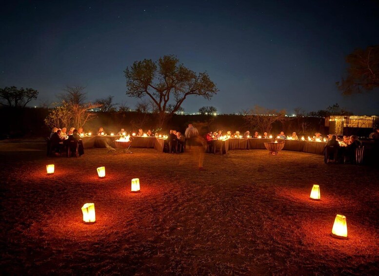 An outdoor gathering with people seated at tables, surrounded by lit candles and lanterns, under a night sky. 1