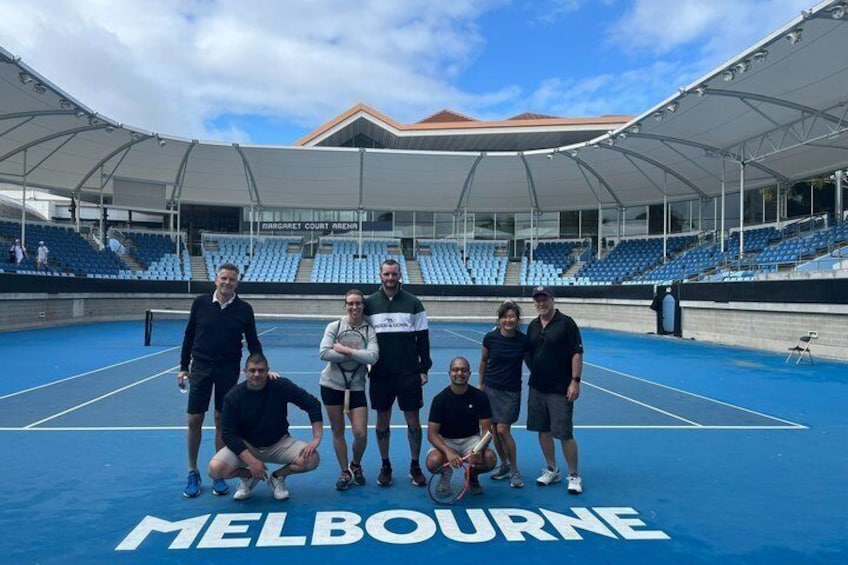 A tennis court with 'MELBOURNE' written on the ground, surrounded by empty blue seats and a covered spectator area. 1