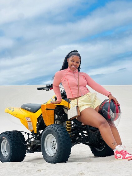 A person sitting on a yellow ATV with a black helmet, wearing pink and yellow clothing, in a sandy area with a cloudy sky. 3