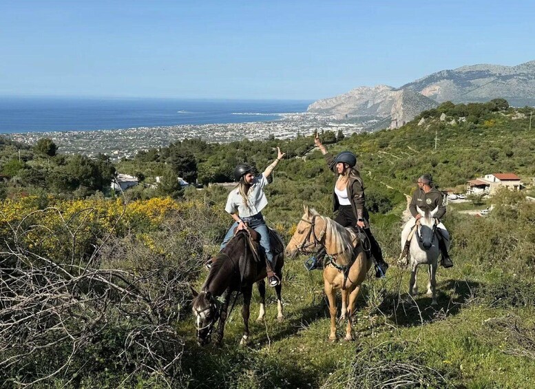 Three people horseback riding on a hillside with a coastal town and mountains in the background. 1