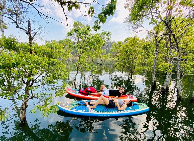 Two people lying on a floating mat in a calm water body surrounded by trees. 1