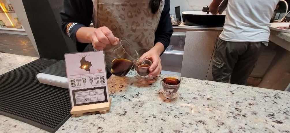 A person pouring a liquid into a glass, with a menu and a bottle on the counter. 4
