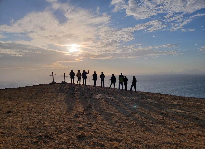 A group of people standing on a hill with a cross, overlooking the sea at sunset. 5