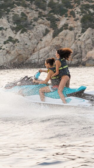 Two people jet-skiing on a body of water with rocky terrain in the background. 1
