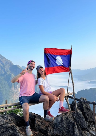 Two people sitting on a rocky outcrop with a flag, mountains in the background. 2