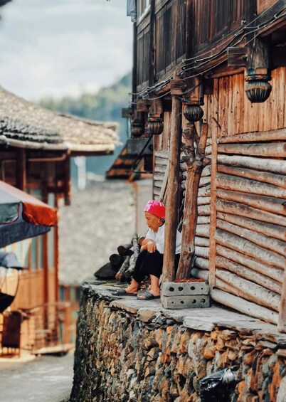 A rustic wooden building with a stone wall, a person sitting on the edge, and traditional architecture in the background. 4