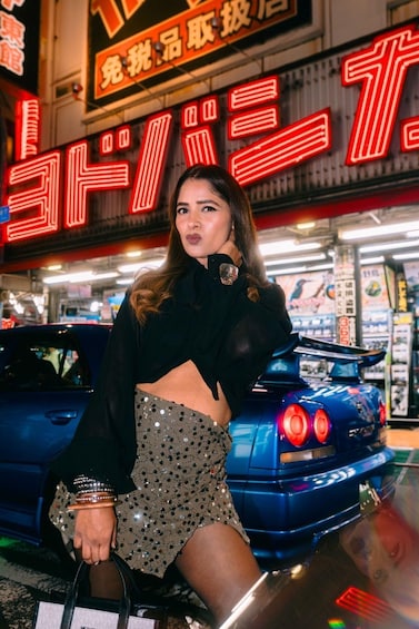A person posing in front of a blue car with a neon sign in the background. 5