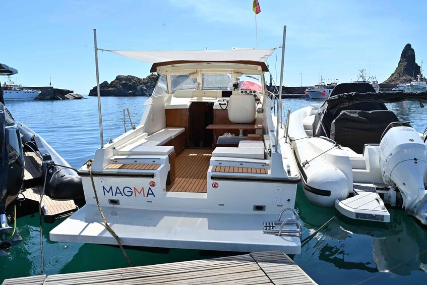 A small white boat named MAGMA docked at a marina with calm water and rocky outcrops in the background. 2