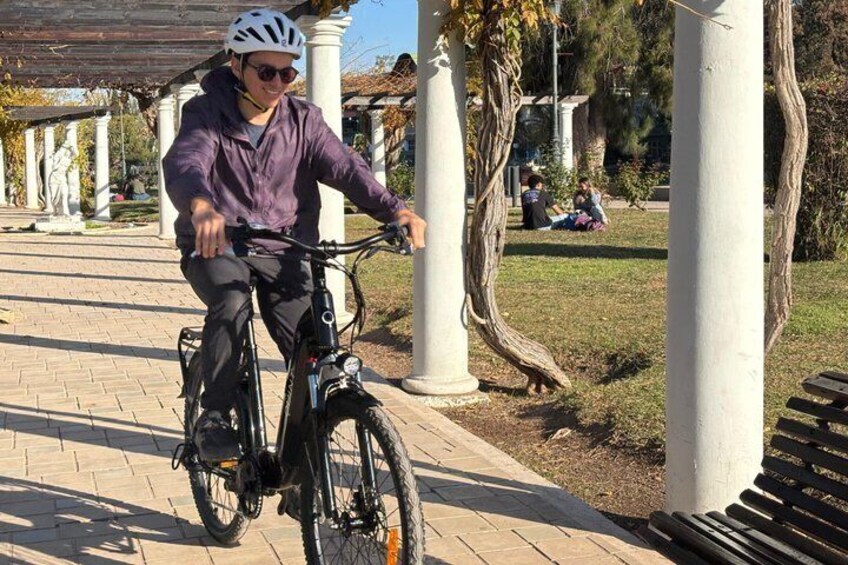 A person riding a bicycle on a paved path with white columns and trees in the background. 3