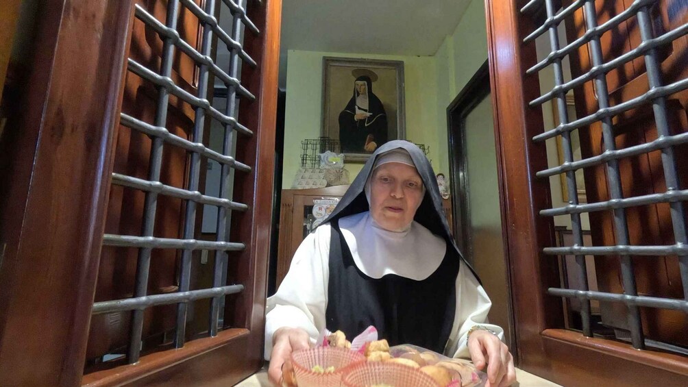 A nun holding a basket of bread in a room with barred windows and a religious painting on the wall. 3
