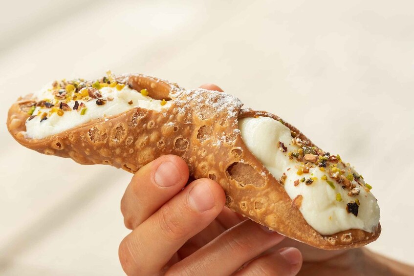 A hand holding a cannoli with white cream filling and chopped nuts. 1