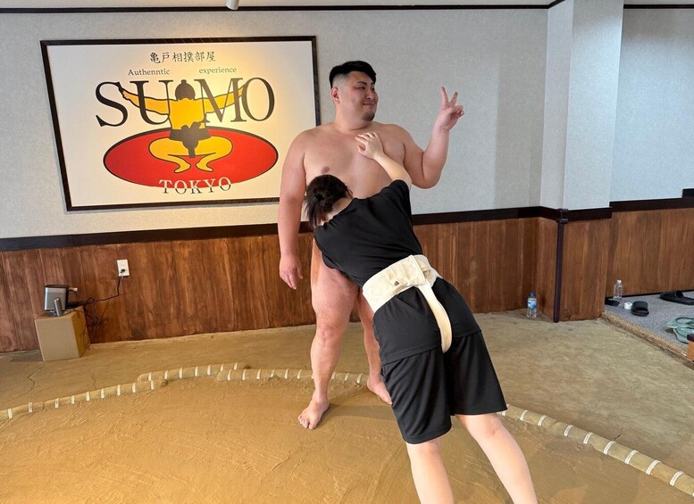 A sumo wrestler in a black outfit with a belt, standing in a room with a sumo wrestling poster on the wall. 4