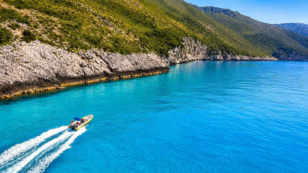 A boat speeding across a clear blue sea with rocky cliffs and green hills in the background. 3