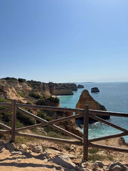 A coastal landscape with rocky cliffs, a wooden fence, and clear blue sea. 3