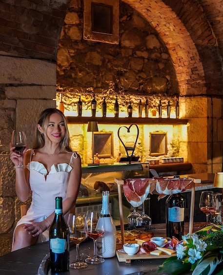 A woman holding a wine glass, a bottle of wine, and a tray of food and cheese in a rustic setting with a brick wall and wine bottles in the background. 5